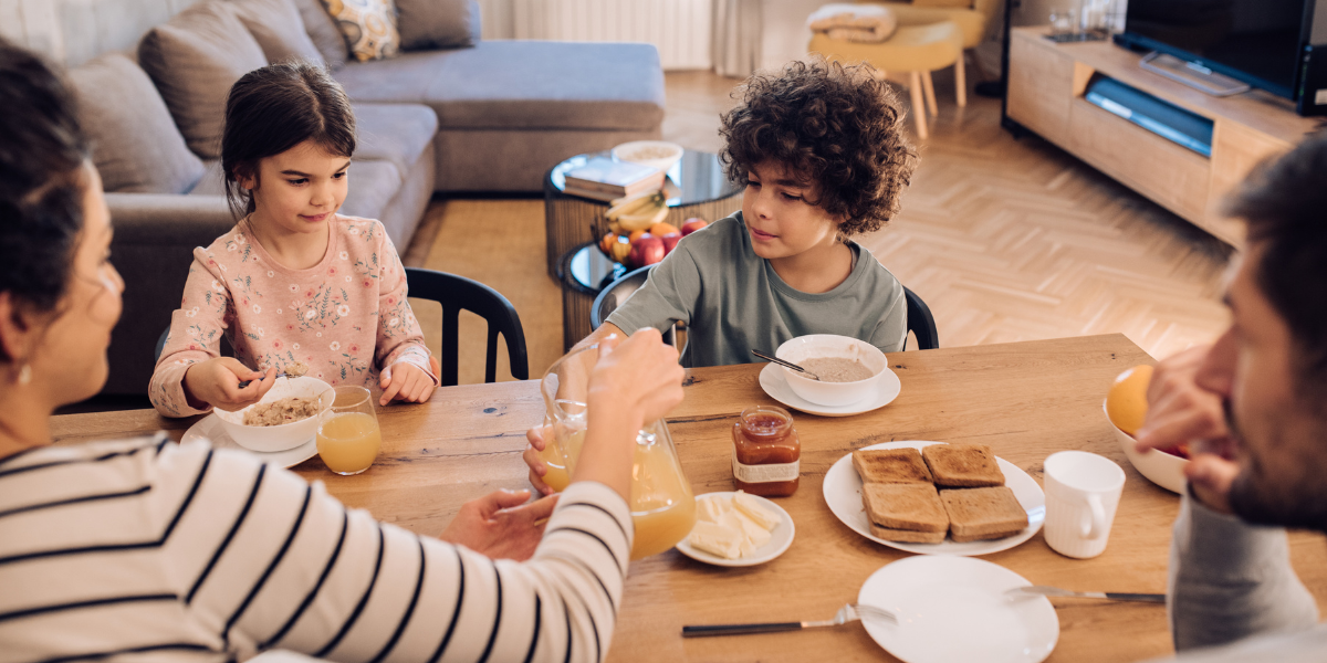 A family of four sits at a table eating breakfast and drinking juice.