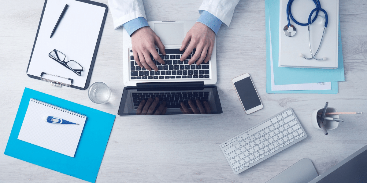 A doctor types on a laptop, a thermometer and stethoscope at their side